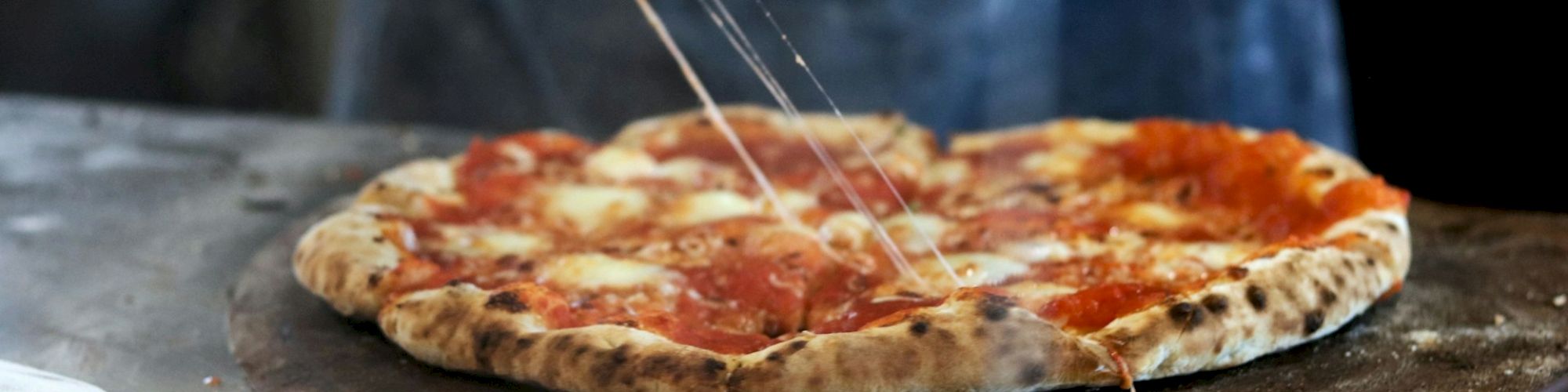 A hand is slicing a freshly baked pizza with cheese, tomato sauce, and pepperoni on a wooden board, with melted cheese stretching.