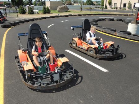 Two children are driving orange go-karts on an outdoor track, with tires lining the course. The track has a clear sunny day backdrop.