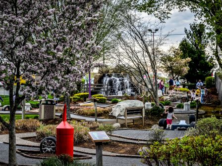 People enjoying a landscaped park with flowering trees, pathways, a red sculpture, and waterfalls in the background.