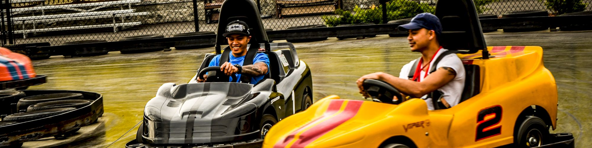 Two people are driving go-karts on a track, with one in a yellow kart and the other in a black kart, at an outdoor amusement park.