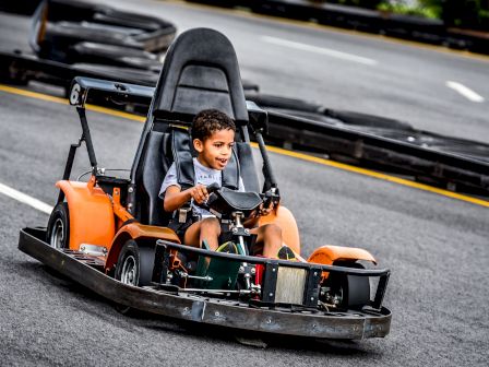 A young child is driving an orange go-kart on a track, appearing happy and focused on navigating the course.