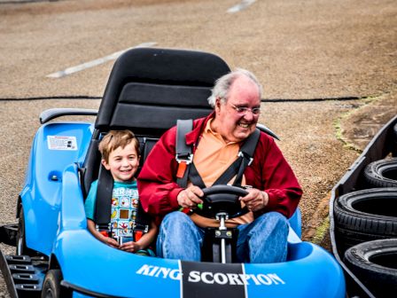 A man and a child are happily riding in a blue go-kart labeled 