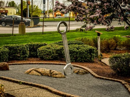 A large wrench sculpture embedded in a landscaped garden area with a rock pathway, bushes, trees, and a fence in the background.