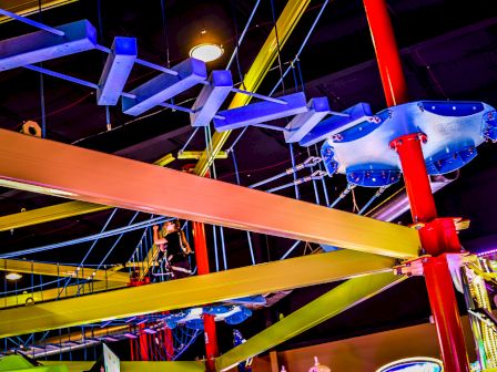 The image shows an indoor ropes course with colorful beams and platforms, along with various obstacles suspended in the air.