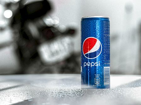 The image shows a blue can of Pepsi placed on a wet surface with a blurred and out-of-focus background.