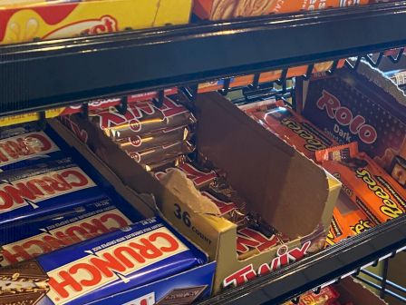 The image shows a display of various candy bars on store shelves, including Crunch, Twix, Rolo, and Reese's.