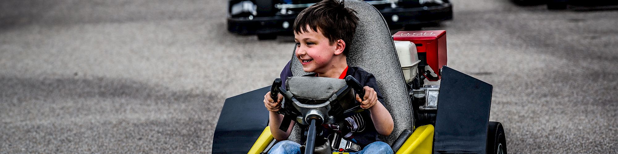 Children driving go-karts on a track, with a young boy in the closest kart smiling and looking ahead, enjoying the activity.