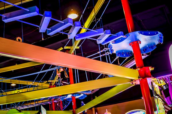 The image shows an indoor ropes course with colorful beams, ropes, and platforms, part of an adventure or amusement area.