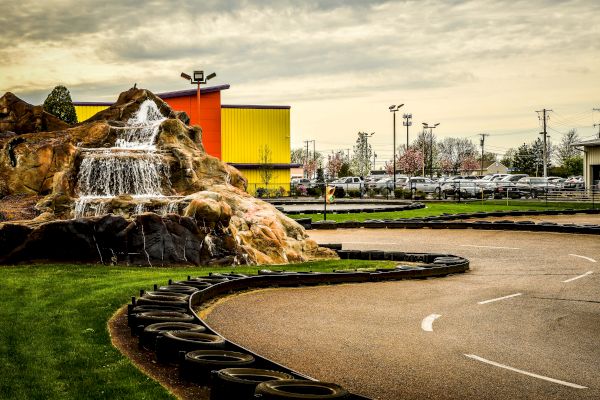 A go-kart track with a curve bordered by tires, adjacent to a rock formation with a waterfall, near buildings and trees under a cloudy sky.