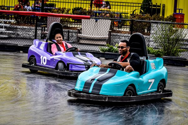 Two people are enjoying a ride in mini electric cars at a fun amusement park attraction, with big smiles on their faces as they drive around.