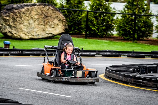 A child is driving a go-kart on an outdoor track, smiling and having fun, while wearing a safety harness and surrounded by greenery and tires.