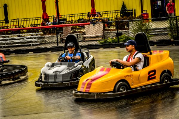 Two people are driving go-karts on a track, one in a gray kart and the other in a yellow kart numbered 2.