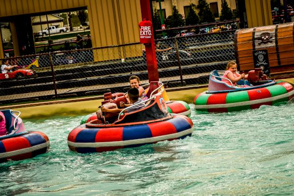 People are riding colorful bumper boats in a pool at an amusement park or similar setting, with water splashing around them.