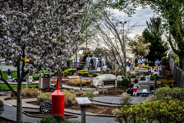 An outdoor scene depicts a park with people strolling, blooming trees, a red structure, pathways, rocks, and a waterfall in the background.