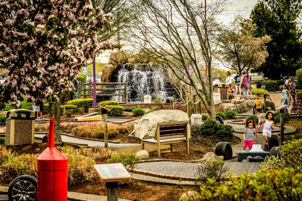 A park scene with children playing and adults walking near a fountain, surrounded by trees, benches, and greenery, and featuring a red buoy decoration.