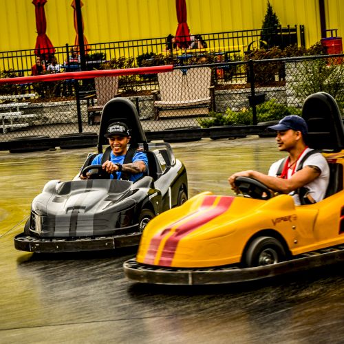 The image shows two individuals driving go-karts, one in a gray car and the other in a yellow car with the number 2, on a go-kart track.