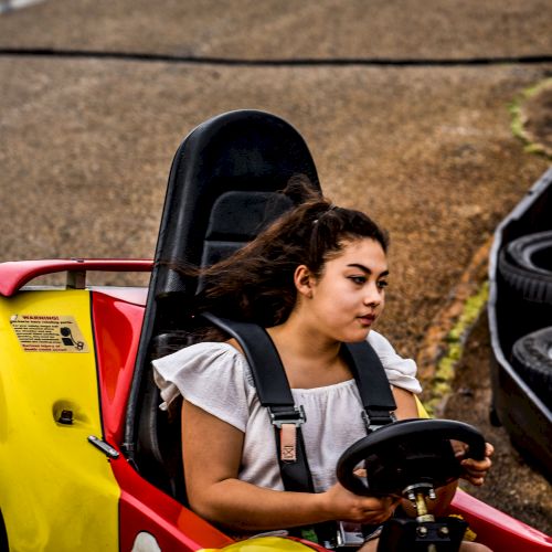 A person is driving a bright yellow and red go-kart on a track lined with stacked tires for safety, concentrating on steering the vehicle.