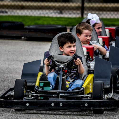 Three children are driving go-karts, with the child in the yellow kart leading the others.