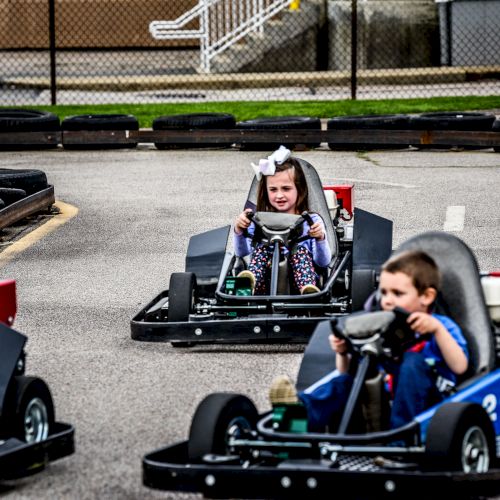 Children are driving go-karts on a track, with a girl in a bow and a boy in blue leading the race, smiles on their faces.