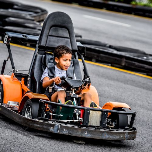 A young child is enjoying a ride in an orange go-kart on a track. The child is smiling and holding onto the steering wheel of the go-kart.