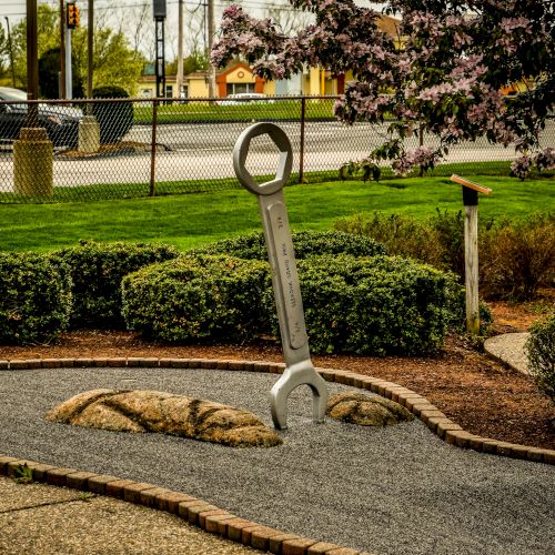 A large wrench sculpture is installed vertically in a landscaped garden with rocks, surrounded by greenery and near a fence and a road.