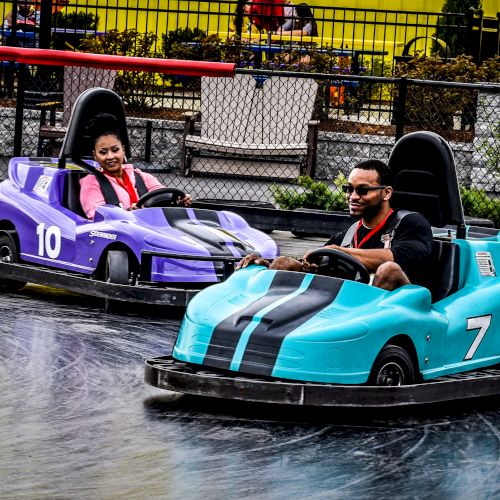 Two people are riding in bumper cars, one in a purple car and the other in a blue car, on an amusement park track.