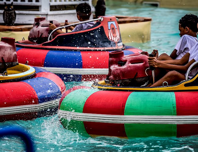 People are riding and bumping into each other with colorful bumper boats in a water ride, splashing water around.