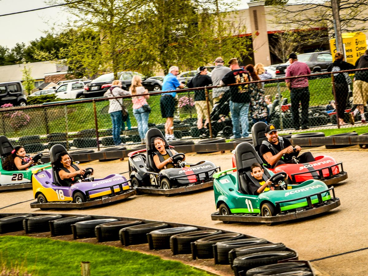 People are driving go-karts on a track, while spectators stand behind a fence watching. The scene is outdoors with trees and parked cars in the background.