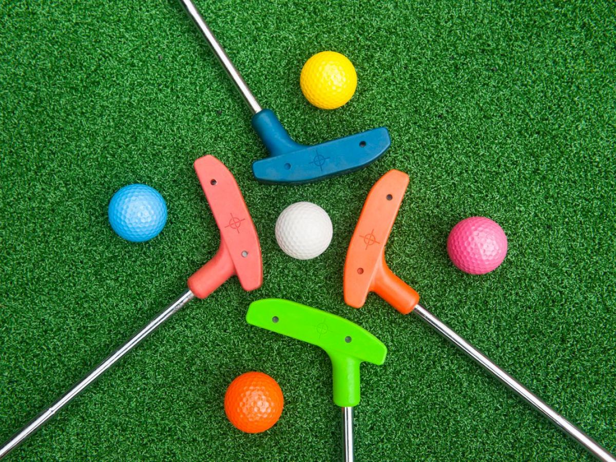 The image shows colorful mini-golf putters and balls arranged in a circle on a green surface, commonly found in miniature golf courses.