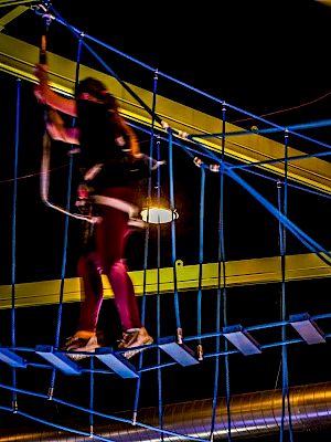 A person is navigating a high ropes course with harnesses and safety gear in what appears to be an indoor adventure park.